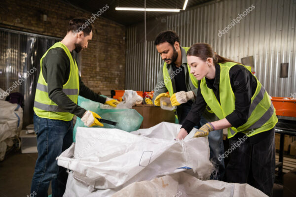 Multiethnic workers in safety vests and protective gloves separating garbage near sacks while working together in waste disposal station, garbage sorting and recycling concept